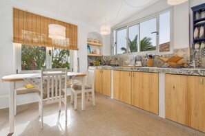 A kitchen with wooden cabinets, a dining table with chairs, a window with blinds, and a shelf with kitchenware.