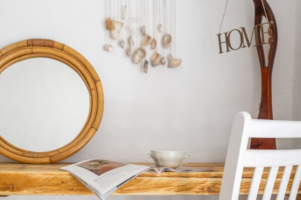 A wooden table with a circular mirror, a white chair, a decorative wall hanging, and a cup.