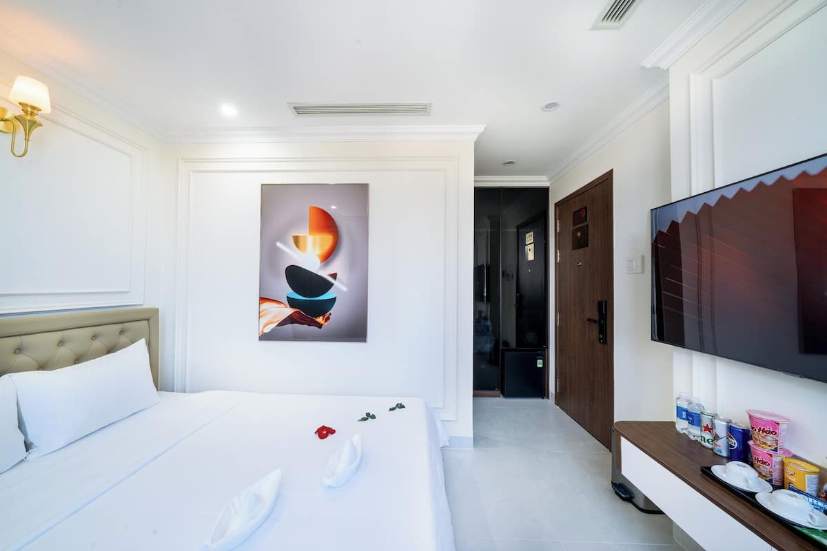 A hotel room with a bed, a wall-mounted TV, a painting, and a small kitchenette area.