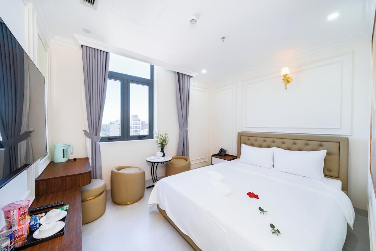 A hotel room with a large bed, a desk with a kettle, a small table with a vase, and a window with curtains.
