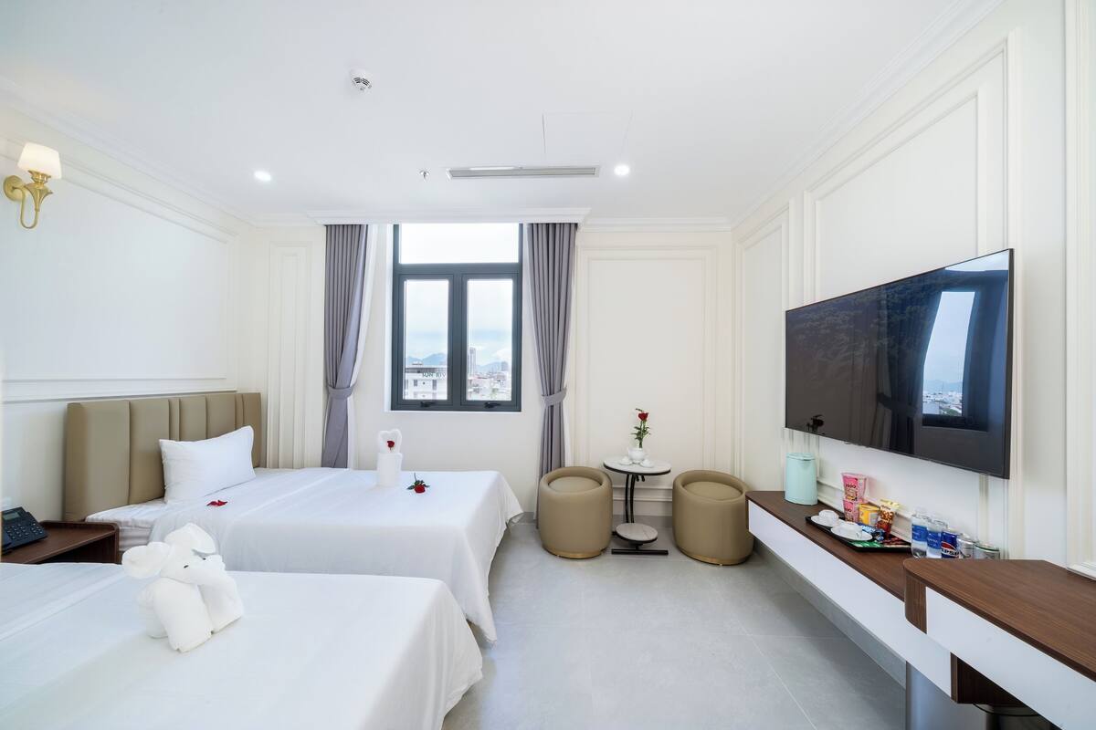 A hotel room with two beds, a TV, a small table with a vase, and a view of the city through the window.