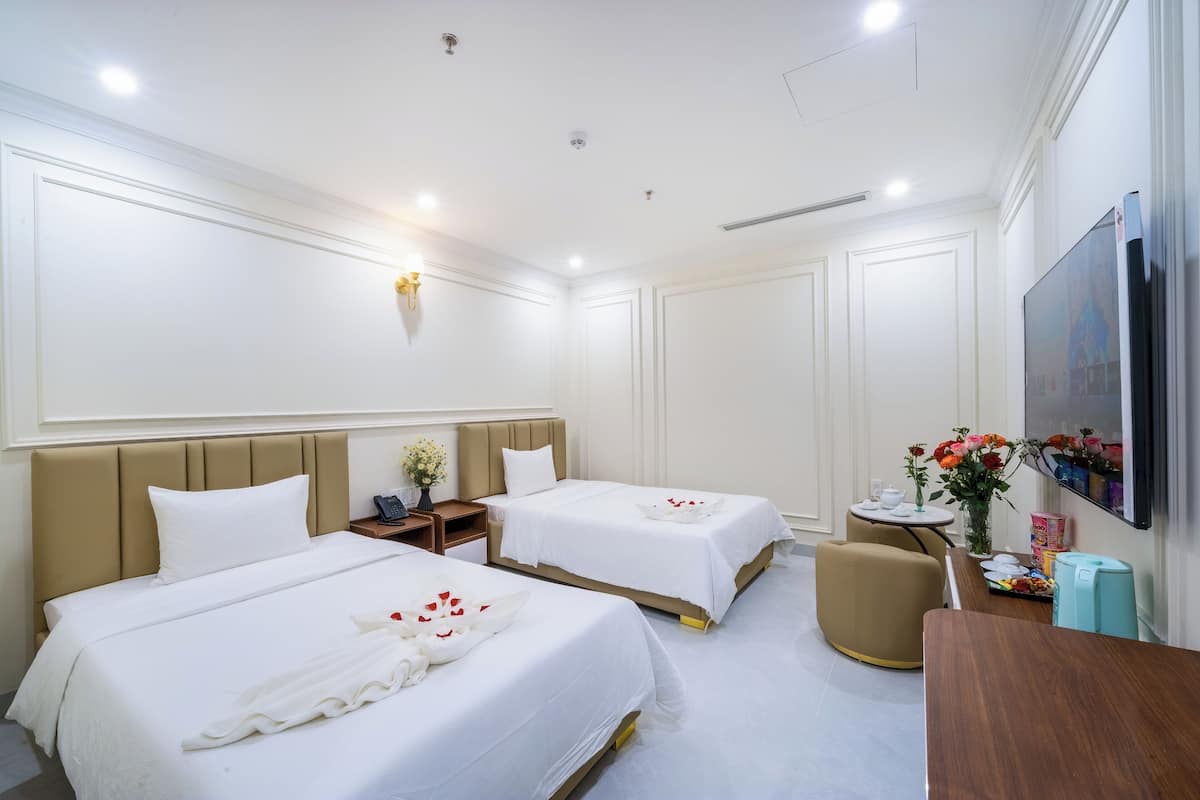 A hotel room with two beds, a desk, a TV, and a small table with flowers.