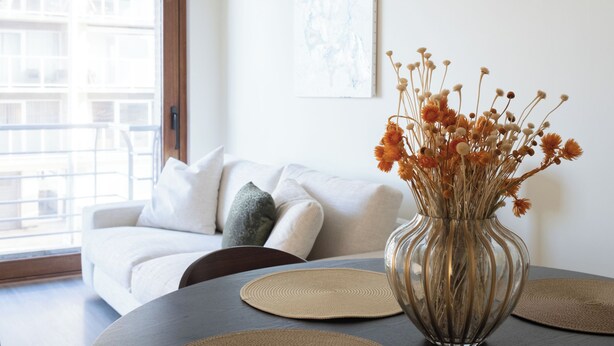 A living room with a white sofa, a glass vase with dried flowers, and a colorful abstract painting on the wall. - Let's Go Raversijde (Oostende)