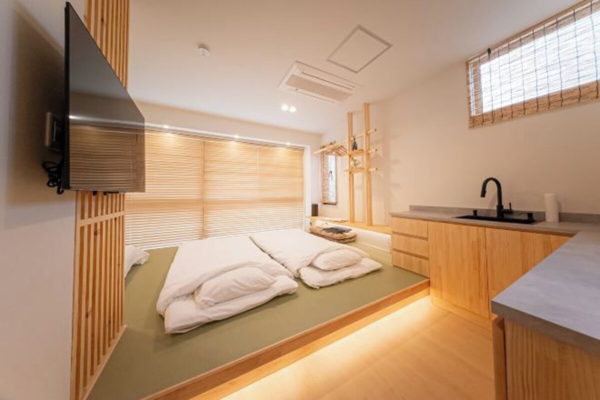 A compact room with a bed, a small kitchenette, and a TV mounted on the wall.
