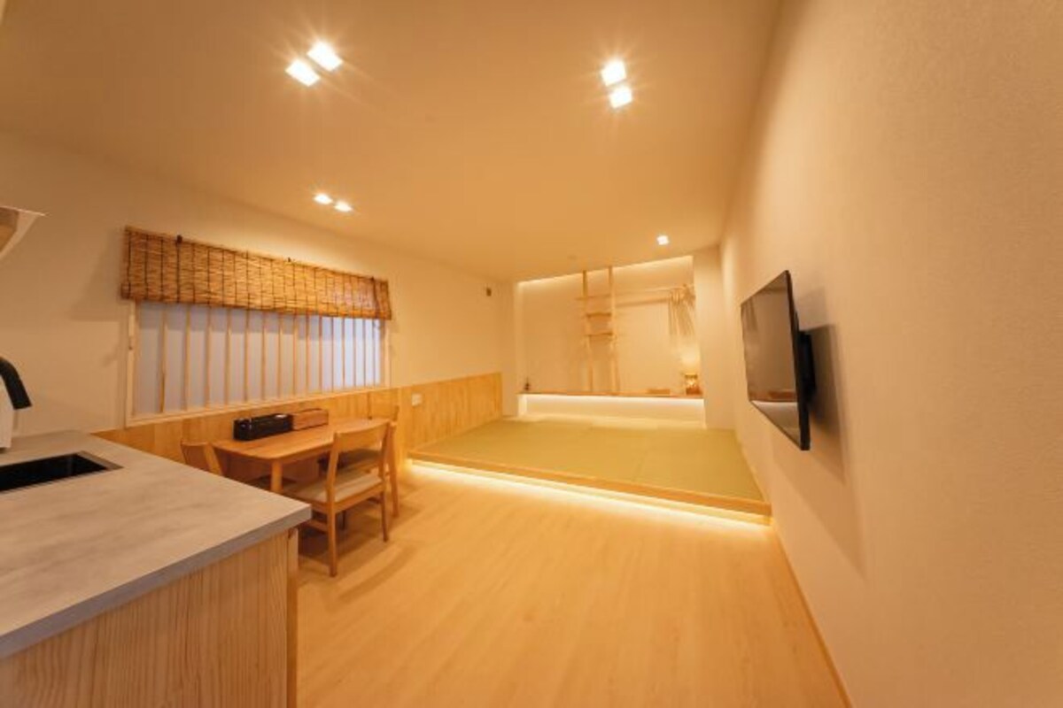 A room with a wooden floor, a dining table with chairs, a kitchen area with a sink, and a television mounted on the wall.