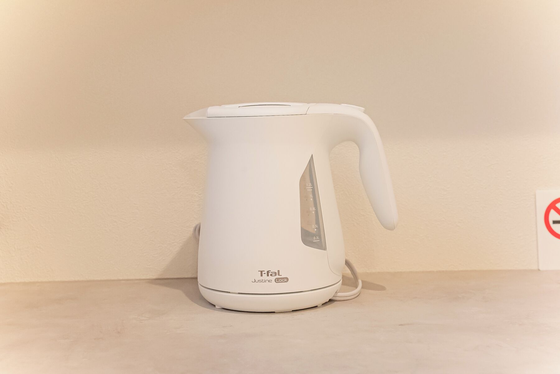 A white electric kettle with a handle and a power cord, placed on a flat surface against a plain wall.