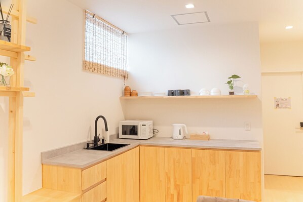 A compact kitchen with wooden cabinets, a microwave, a kettle, and a small shelf with kitchenware.