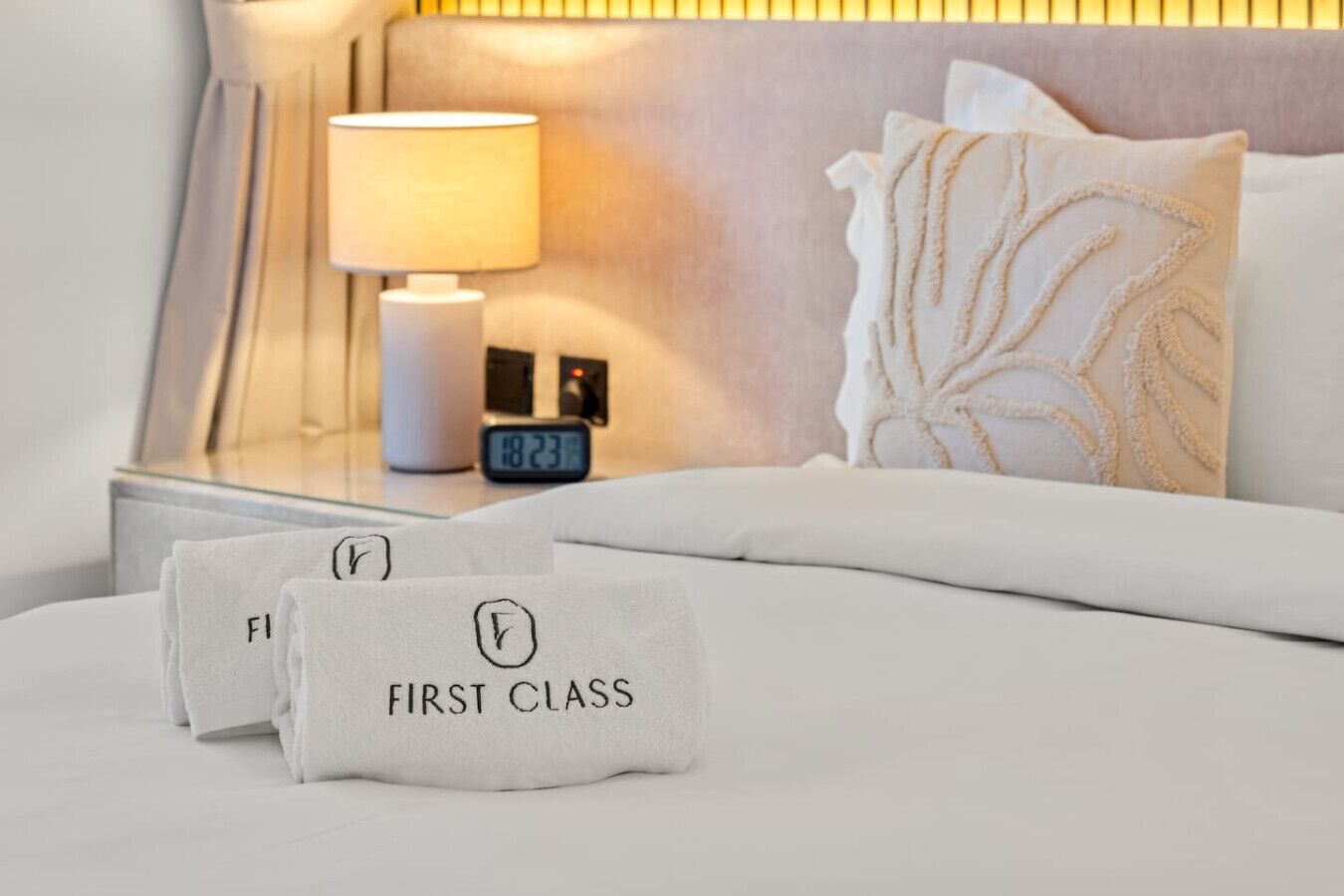 Una cama bien hecha con ropa blanca, un almohadón con estampado de hojas, una lámpara de noche y un reloj digital que marca las 12:23.