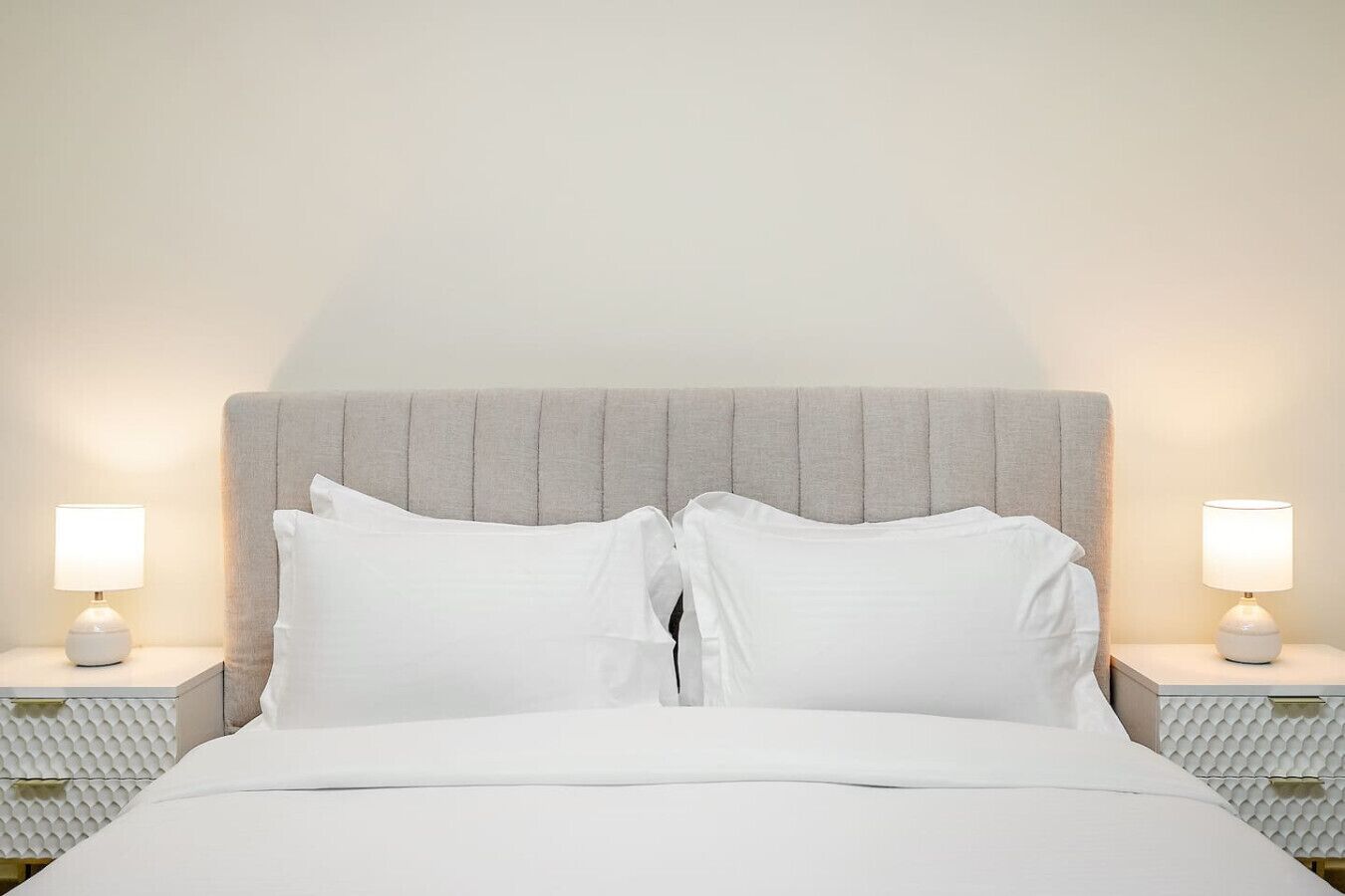 A neatly made bed with white linens and pillows, flanked by two bedside tables with lamps, set against a plain wall.