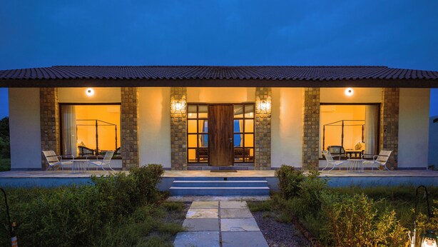 A two-story house with a stone facade, a wooden door, and two outdoor seating areas with chairs and tables. - Nature Jawai Retreat | Luxury Private Pool Villa Resort, Jawai (Bali)