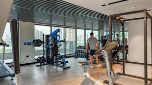 A modern gym with various exercise equipment, large windows offering a city view, and a wooden floor. - FIRST CLASS | Studio | Minimalist Chic Retreat (JVC)