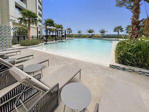 Zona de piscina amb gandules i taules, una piscina nítida i un edifici al fons.