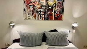 Une chambre d’hôtel avec un lit, des oreillers à rayures et un tableau abstrait aux couleurs vives accroché au mur.