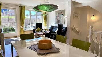 Ein modernes Wohnzimmer mit einem weißen Esstisch, grünen Stühlen, einem schwarzen Sofa und einer grünen Pendelleuchte.