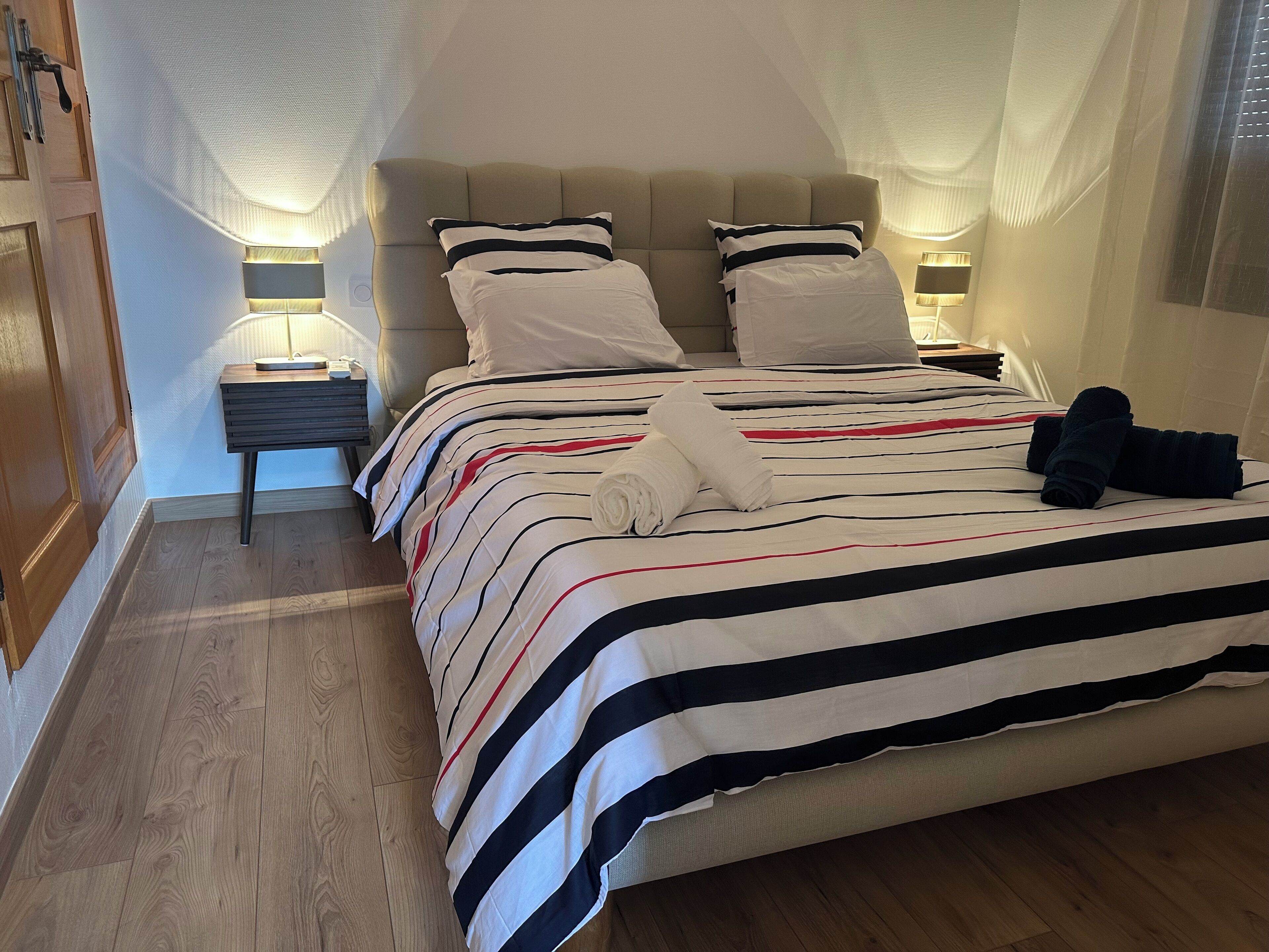 A neatly made bed with striped bedding, two bedside lamps, and a wooden floor.