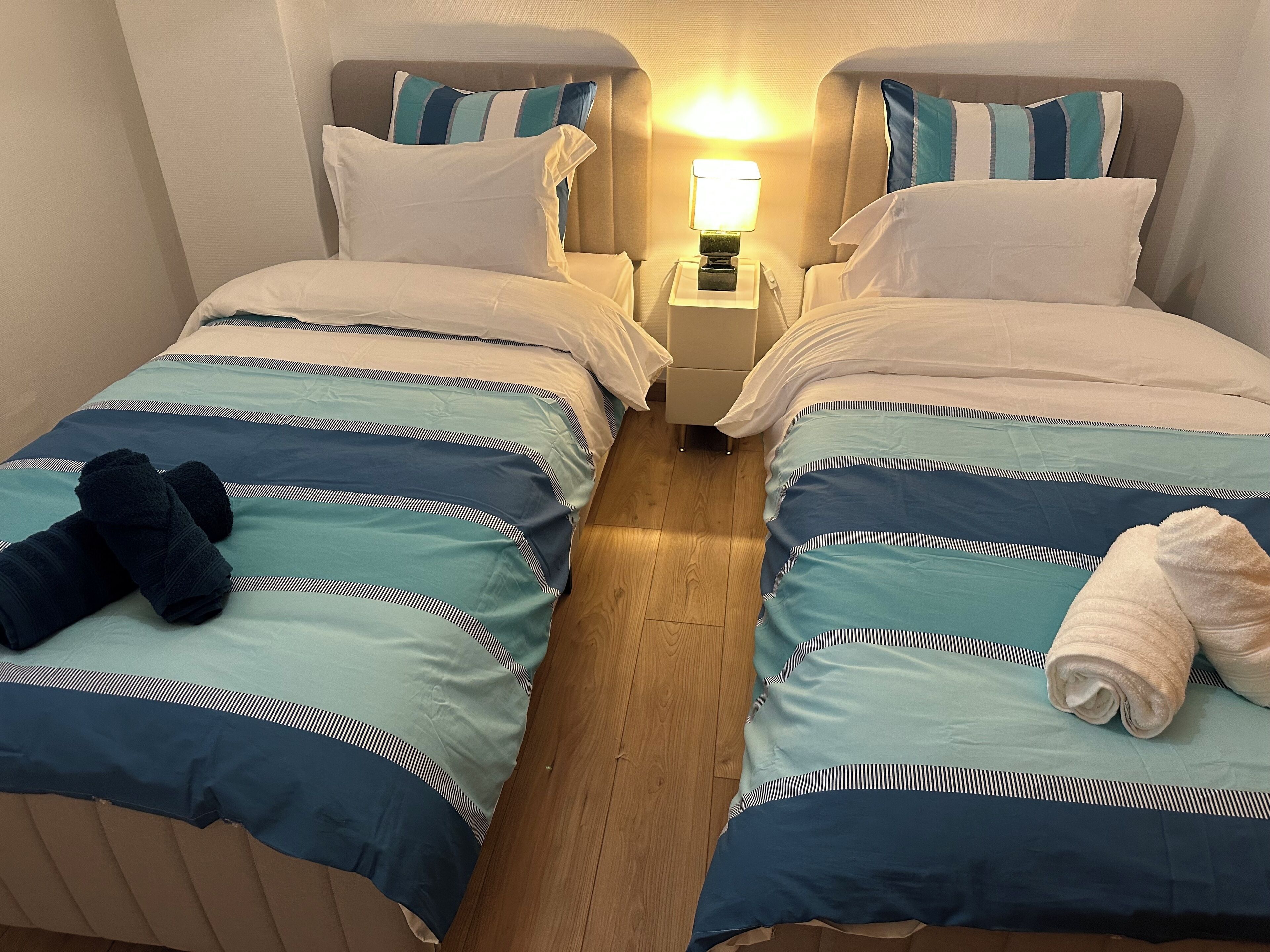 Ein Hotelzimmer mit zwei Betten, jeweils mit einer blau-weißen Streifenauflage, weißen Kissen und einem Nachttisch mit einer Lampe dazwischen.