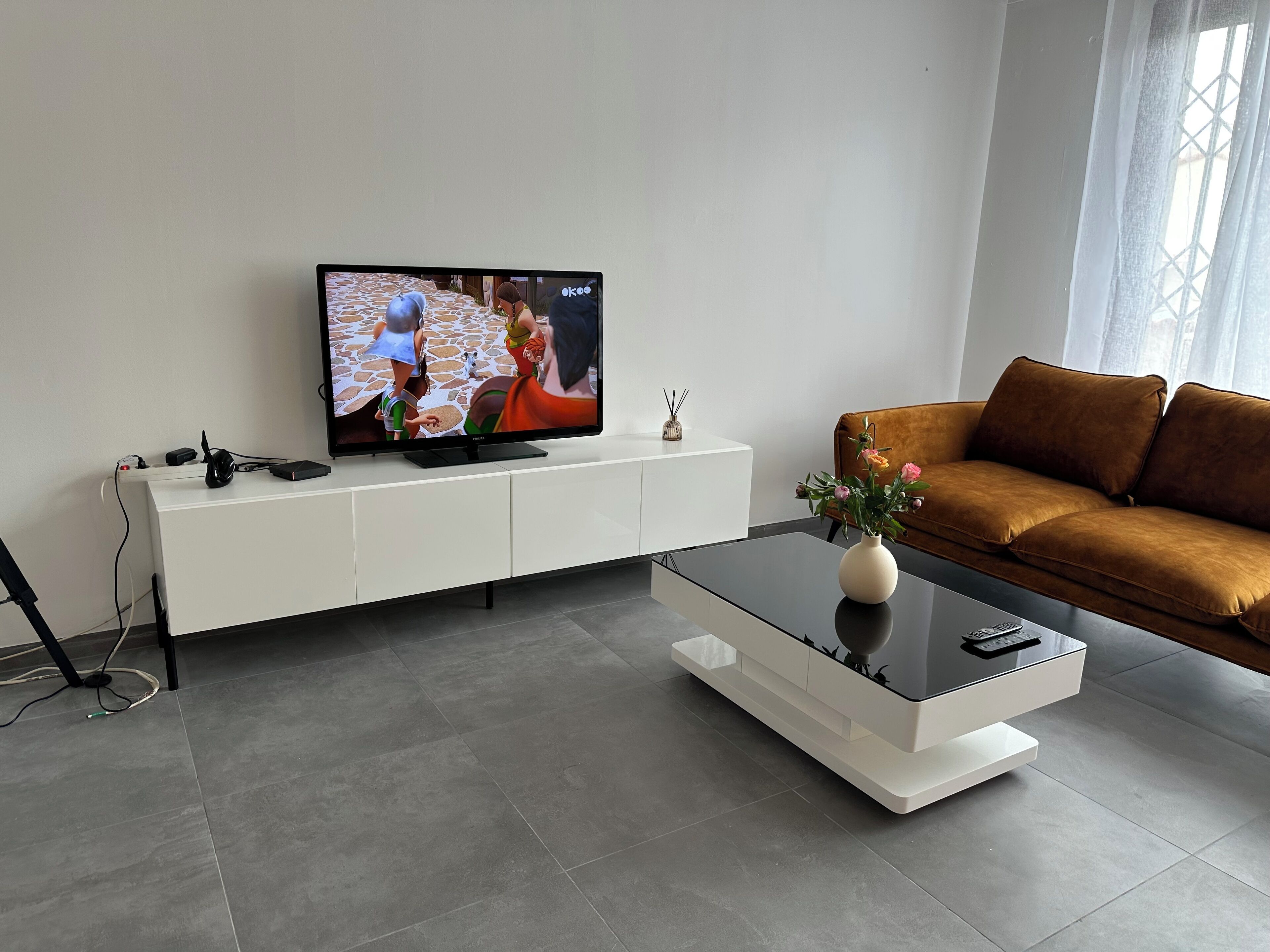 Ett modernt vardagsrum med en platt-TV placerad på ett vitt skåp, en brun soffa och ett soffbord i glas med en vas.
