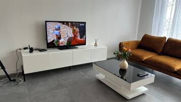 Ett modernt vardagsrum med en platt-TV placerad på ett vitt skåp, en brun soffa och ett soffbord i glas med en vas.