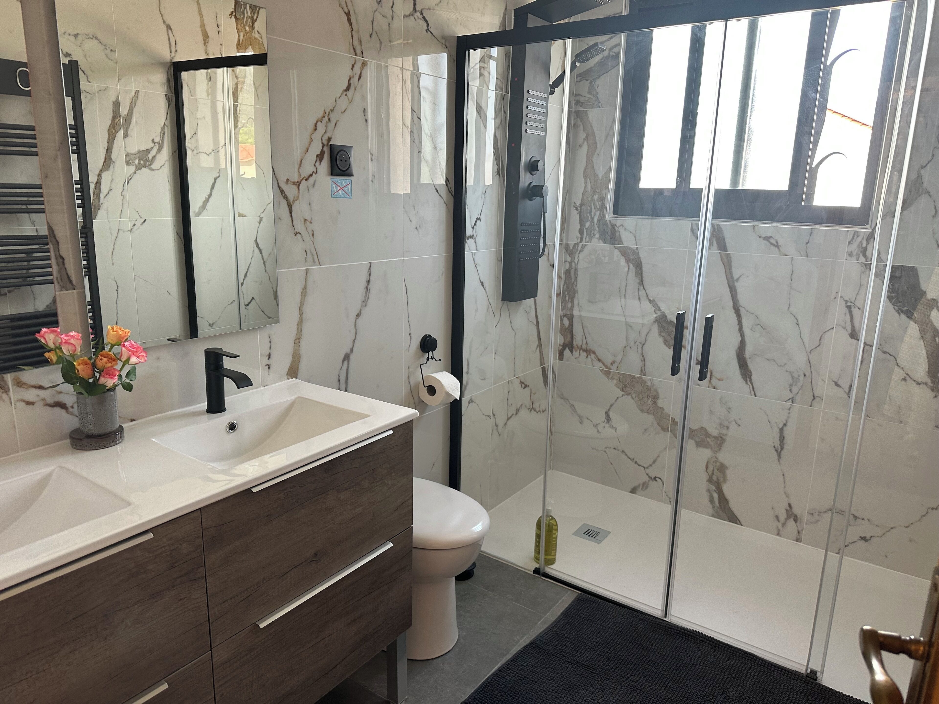 Un baño moderno con un salpicadero de mármol, un lavabo con tocador de madera, inodoro y zona de ducha con mampara de cristal.
