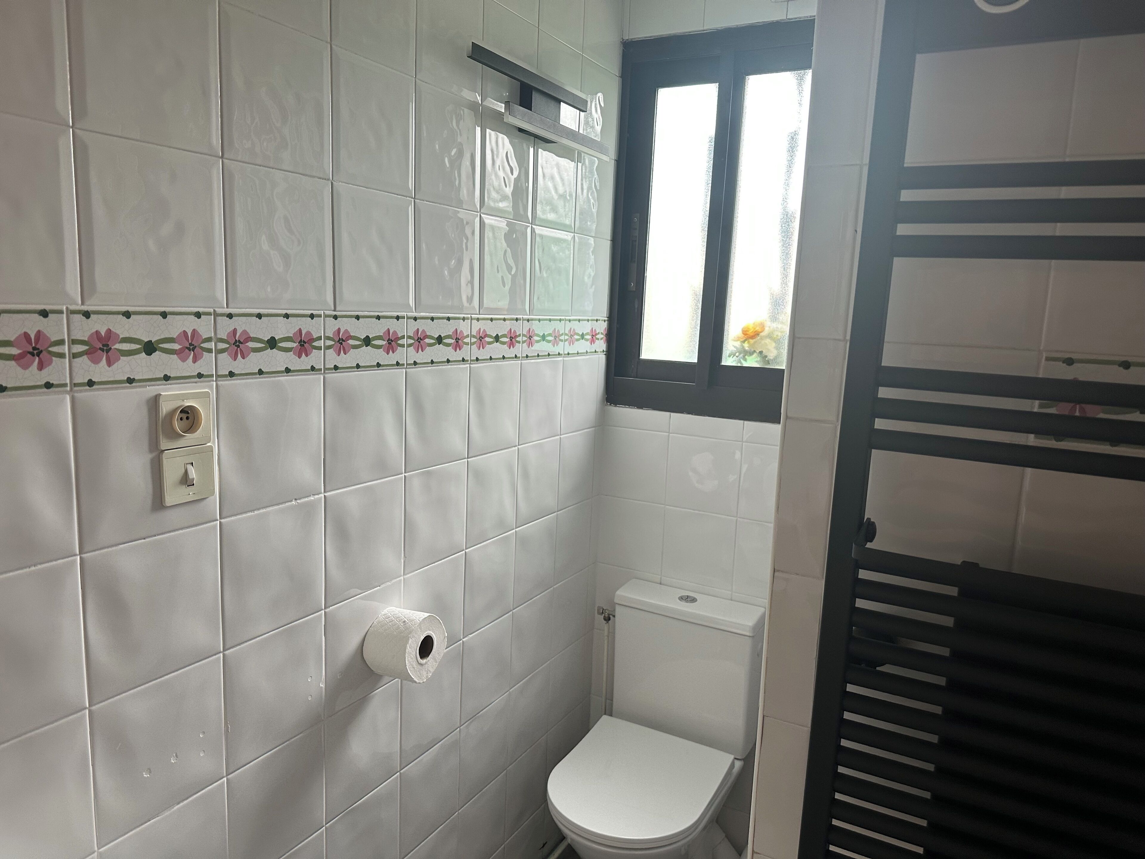 A small, clean bathroom with white tiles, a toilet, a paper towel dispenser, and a window with a view of flowers outside.