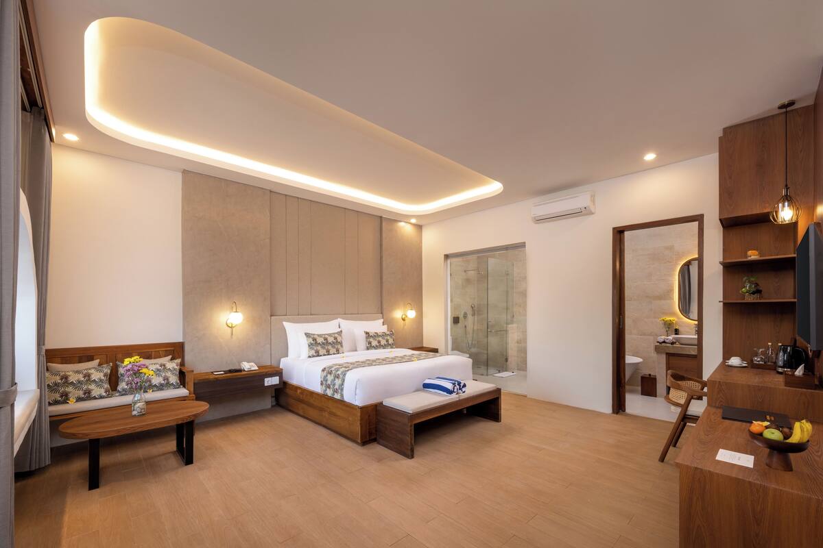 Une chambre d’hôtel moderne dotée d’un grand lit, de tables de chevet, d’un banc en bois, d’un bureau avec une chaise, d’un miroir et d’un espace douche.