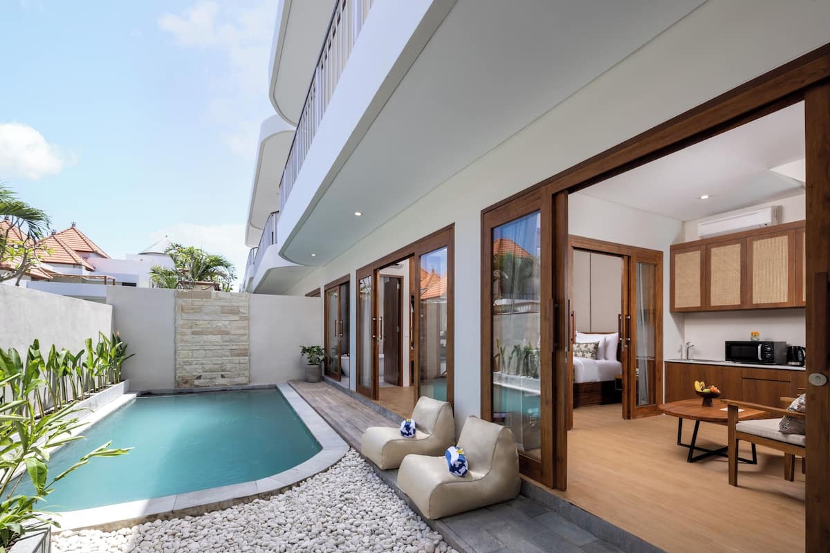 Un espace piscine intérieur moderne, avec une petite piscine, deux chaises longues, une table en bois sur laquelle se trouve un saladier rempli de fruits, et un coin cuisine visible à travers des portes vitrées.