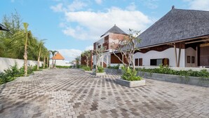 A courtyard with a paved walkway, a building with a tiled roof, and a palm tree. - La Tropica Canggu Bali (Buduk)