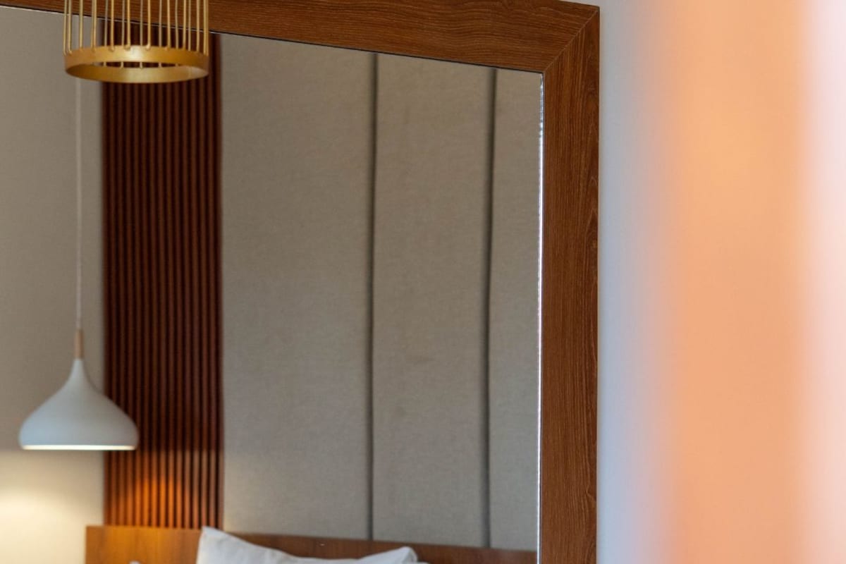 A wooden bedside table with a tissue box and a bowl. A large mirror reflects a bed with pillows and a wall with vertical wooden slats.