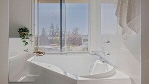 Ein modernes Badezimmer mit einer großen Badewanne, einem Fenster mit Blick auf Bäume und Gebäude sowie einem Blumentopf auf der Fensterbank.