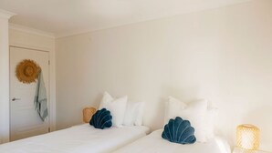 Une chambre avec deux lits, du linge de lit blanc, un chapeau de paille accroché à la porte et un panier tressé posé sur l’un des lits.