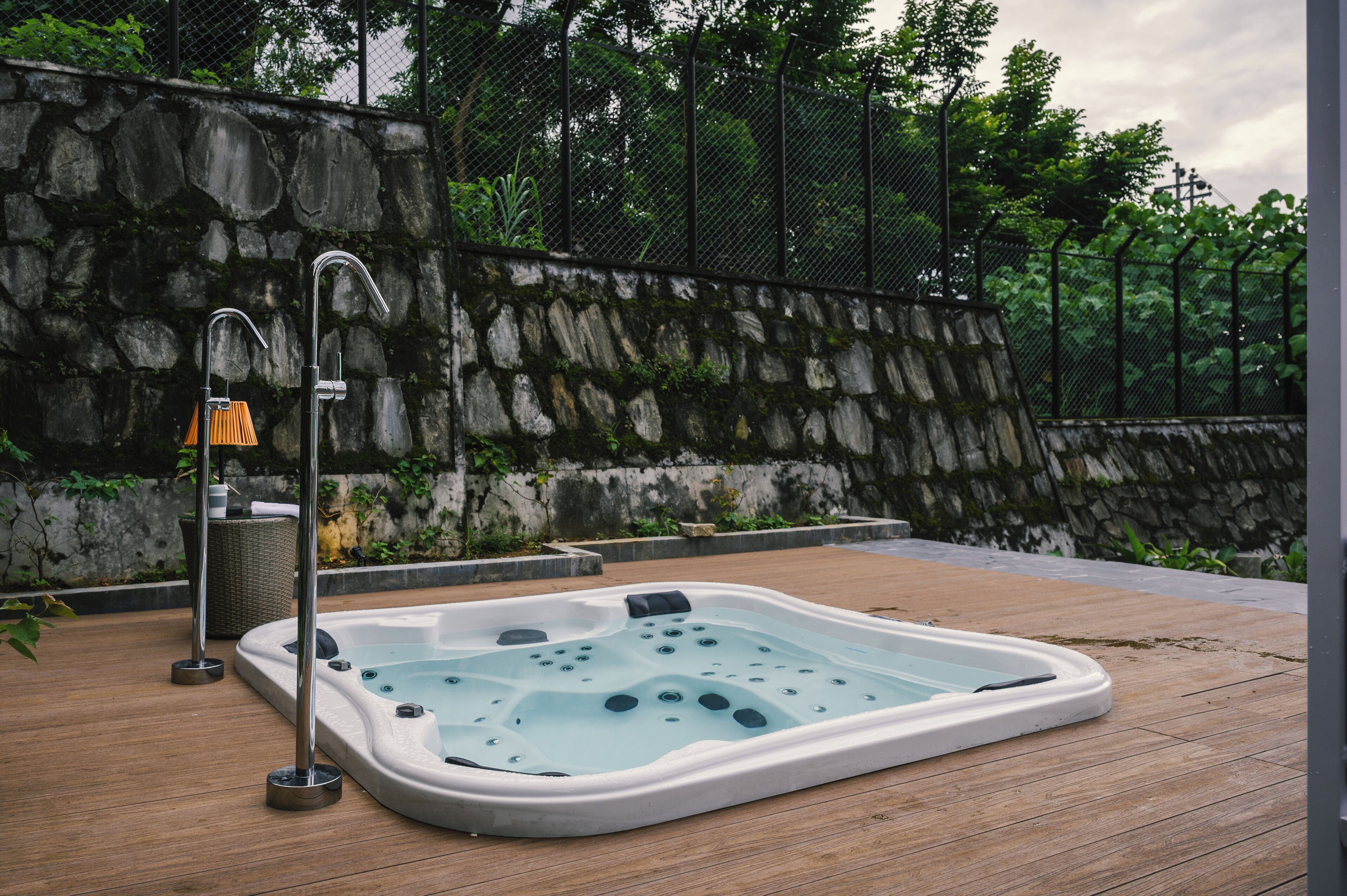 Un jacuzzi en una terraza de madera con un muro de piedra y una valla metálica al fondo.