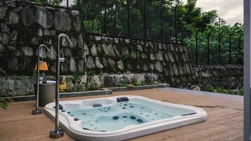 Un jacuzzi en una terraza de madera con un muro de piedra y una valla metálica al fondo.