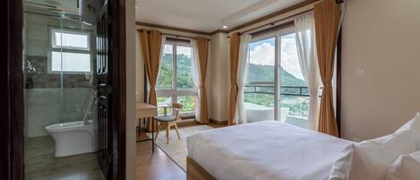 Habitación de hotel con una cama grande, un baño con inodoro y ducha, una mesita con una silla, y un balcón con vistas a las montañas.