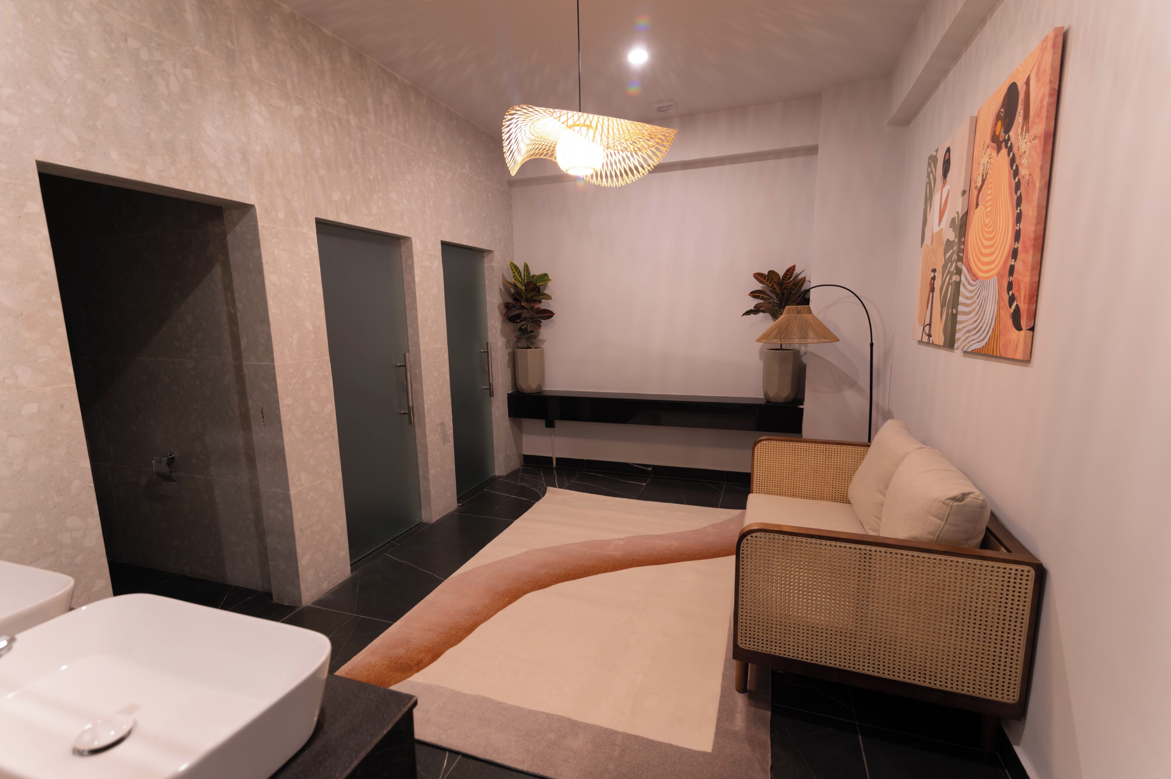 Une salle de bain moderne avec un lavabo blanc, des carreaux de sol noirs et beiges, un tapis beige, un canapé en osier, une lampe sur pied et des œuvres d’art murales.