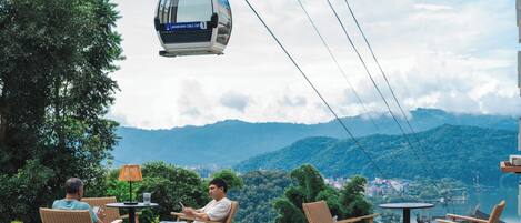 Un teleférico suspendido sobre una terraza con mobiliario de mimbre y vistas a un paisaje montañoso.