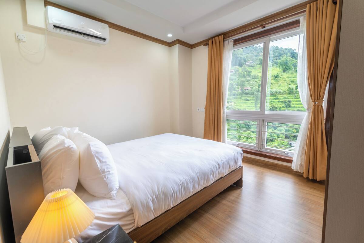 A bedroom with a bed, an air conditioning unit, a lamp, and a window with curtains.