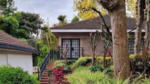 A residential area with a house featuring a tiled roof, a staircase leading to the entrance, and a garden with various plants and shrubs. - Thayu House (Limuru)