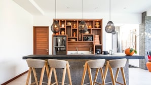 A modern kitchen with a central island, wooden bar stools, hanging pendant lights, and a built-in shelving unit with various kitchen items. - Stunning 6 Bedroom Villa Only 400m to the Beach (Canggu)