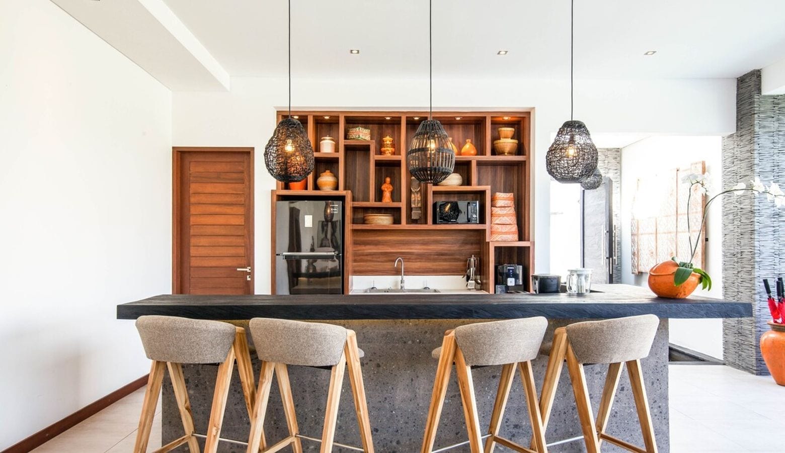 Eine moderne Küche mit einer zentralen Kochinsel, Holzbarhocker, hängenden Pendelleuchten und einer eingebauten Regaleinheit mit diversen Küchenutensilien.