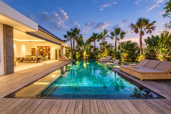 A modern outdoor pool area with a wooden deck, lounge chairs, and a dining area with a kitchen visible in the background.