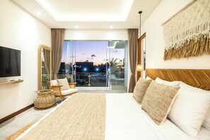 A modern hotel room with a large bed, a television, a balcony with a city view, and a woven rug. - Seaside Luxury: 4BR Villa, 400m to Finns, Canggu (Canggu)