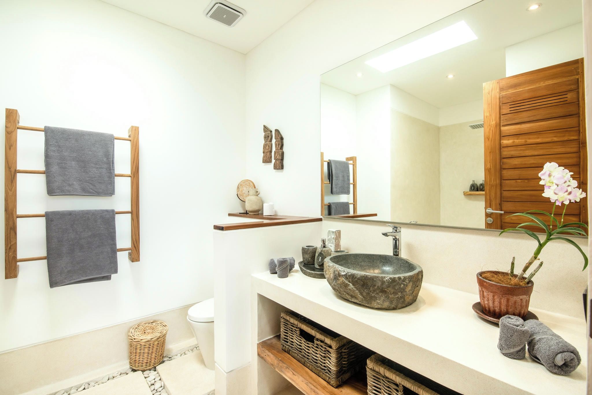 A bathroom with a wooden towel rack, a large mirror, a stone sink, wicker baskets, and a potted plant.