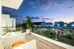 A balcony with wicker furniture, a wooden floor, and a glass railing overlooking a cityscape at dusk. - Seaside Luxury: 4BR Villa, 400m to Finns, Canggu (Canggu)