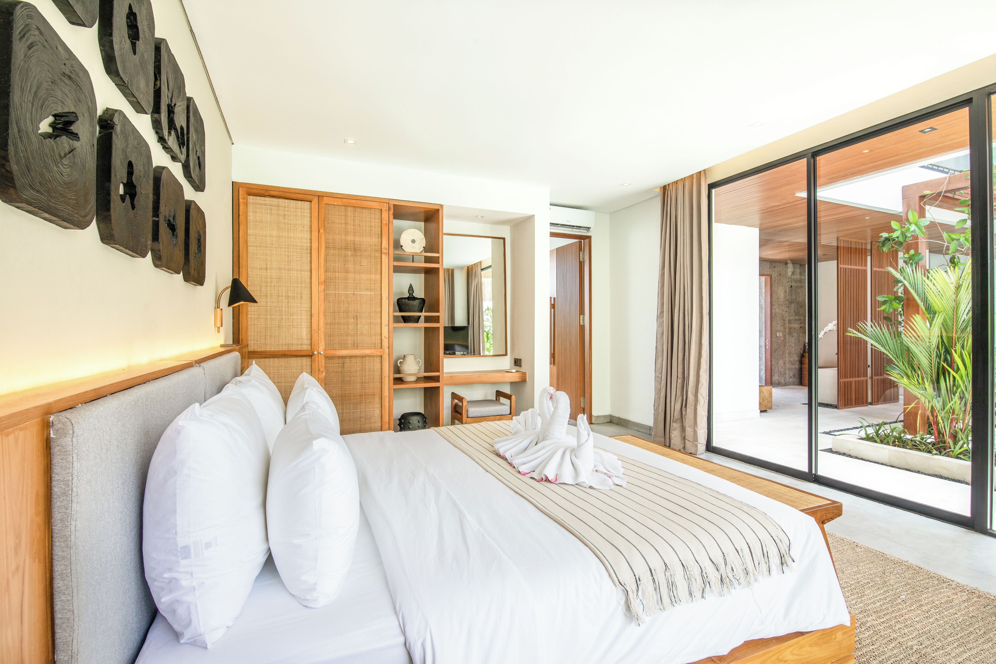 A modern hotel room with a large bed, a wooden wardrobe, a small desk, and a sliding glass door leading to a balcony with plants.