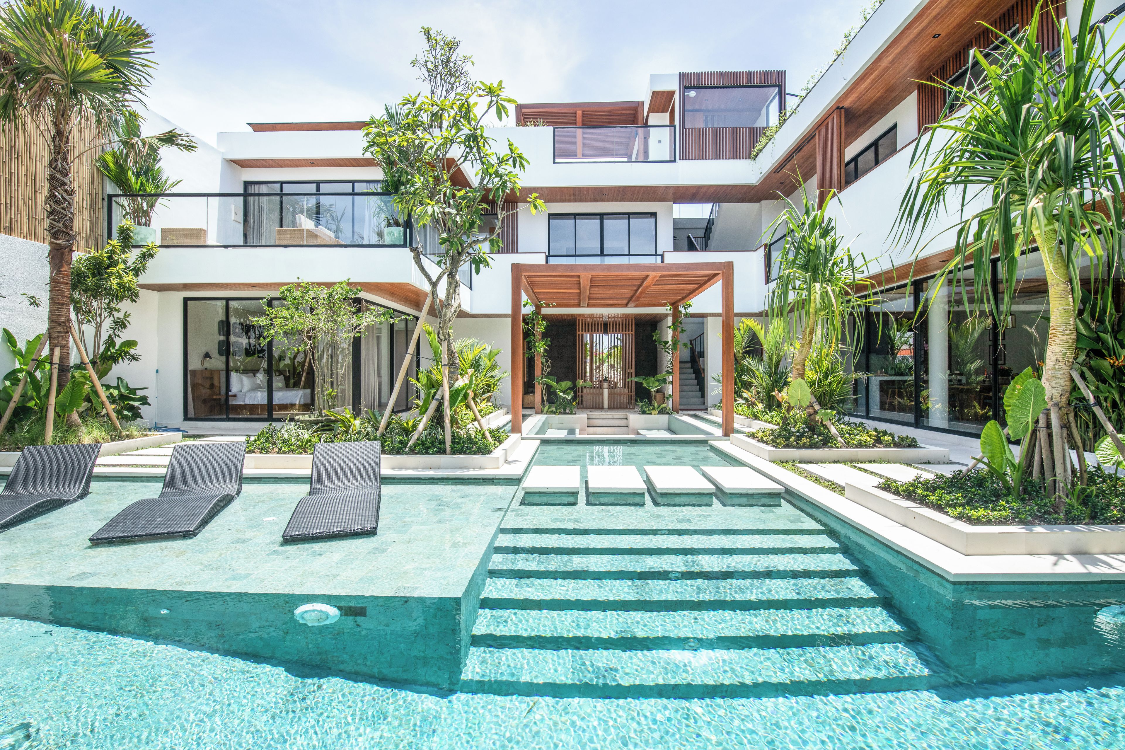 A modern multi-story building with a swimming pool, lounge chairs, and tropical plants.