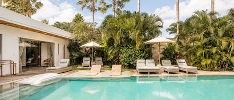 A pool with lounge chairs and umbrellas, surrounded by palm trees and a building.