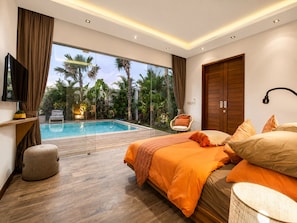Un dormitorio con una cama grande, televisión, suelo de madera, piscina y vistas a palmeras.