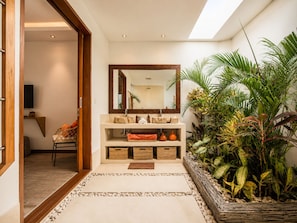 A modern bathroom with a large mirror, a sink, and a shower area. There is a wooden door leading to another room, and a small seating area with a chair and cushions.