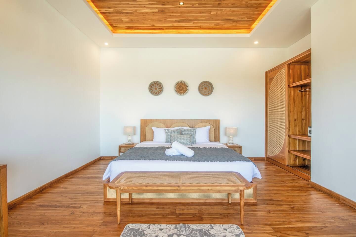 A spacious bedroom with a wooden ceiling, a large bed with a woven headboard, two bedside tables with lamps, and a wooden wardrobe.