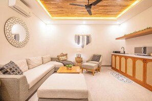 A modern living room with a sofa, a coffee table, a chair, a decorative mirror, a ceiling fan, and a kitchen area with a microwave.